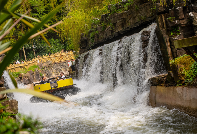 "Boat ride passing under a large waterfall, splashing water around riders in an outdoor theme park."