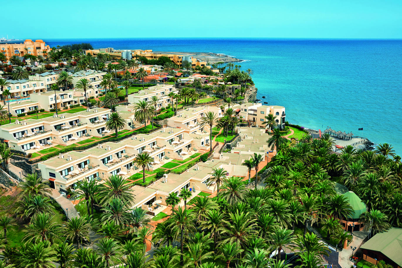 Aerial view of the hotel complex with clusters of rooms one above the other going up the hill with many palm trees and the bottom and the lovely blue sea below.
