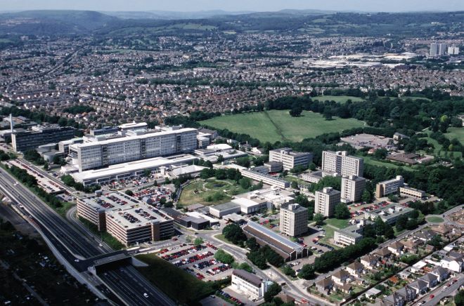 "aerial view of Heath Park campus, large university buildings in the foreground, with rows of houses behind. In the distance green fields turn into hills"