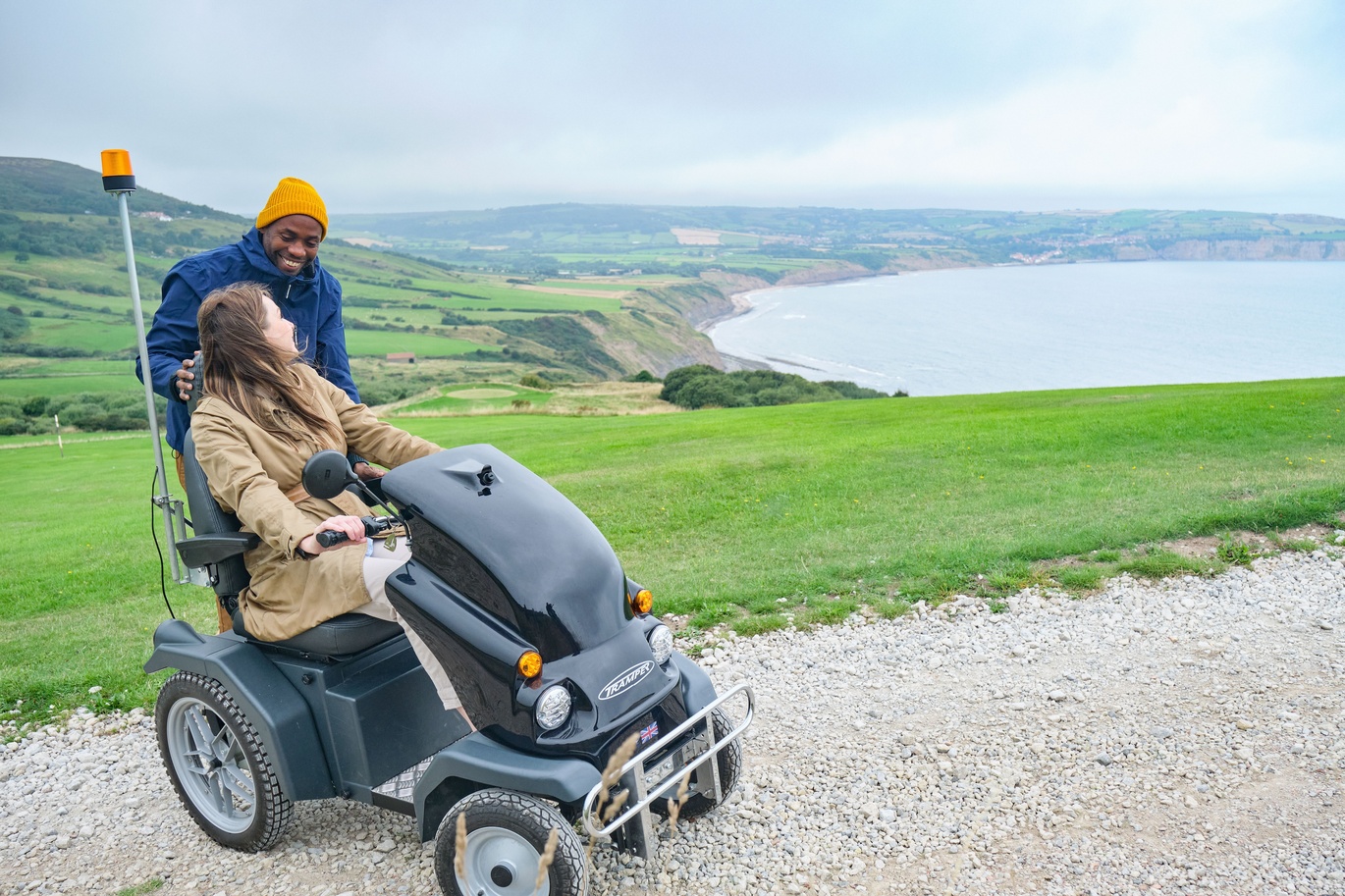 "Male standing next to a female using an electric mobility scooter, next to a scenic view over cliffs overlooking the sea"