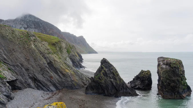 View of the beach and sea with large rocks surrounding and low clouds over the mountains behind.