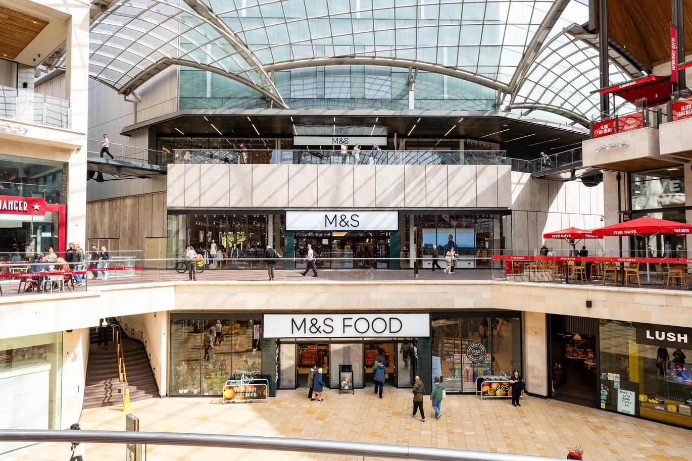 "Interior view of Cabot Circus showing M&S and M&S Food entrances, glass roof, upper walkways and outdoor seating with shoppers."