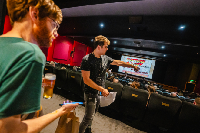 An indoor setting of a movie theater with several individuals seated, looking towards the screen where a colorful title is displayed. Two individuals stand in the foreground; one appears to be gesturing towards the screen, holding papers and a pen, while the other holds a drink. 