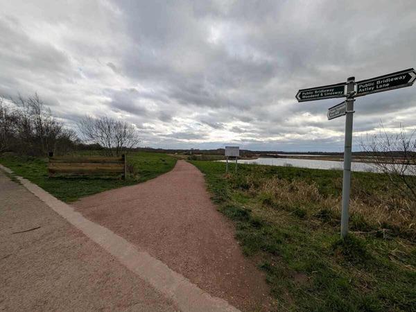 A wide gravel path runs alongside a lake and grassy landscape, with signposts indicating public bridleways under a cloudy sky