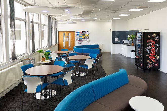 "Student accommodation common room with chairs around tables, and long sofas. A kitchen is in the rear, with a snack machine to the right"