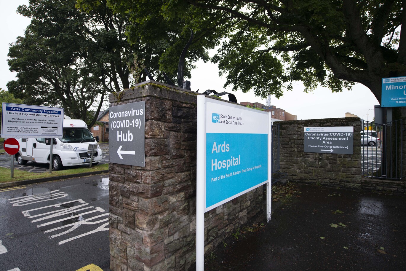 "External signage to Ards Hospital next to a stone wall. There are also car parking signs and signs for the COVID-19 Hub and COVID-19 Priority Assessment Area. Behind the stone wall is a car park and a campervan is parked in one of the bays and there are large oak trees above the wall and car park."