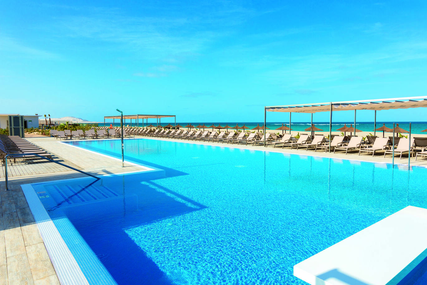 View of the pool surrounded by many sun loungers with the sea in the background on a sunny day.