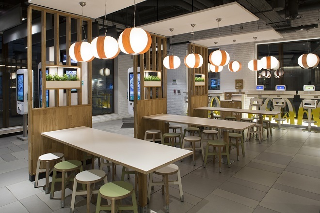 "Photo showing inside of a McDonald's restaurant. There are green and white stool seats around long rectangular tables, seating 8 per table. There are self service checkout screens near the window to the left. There are white and orange circular lights hanging from the ceiling."