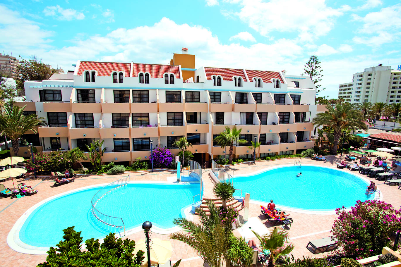 Aerial view of the pool area with a bridge over the pool and people around on sun loungers. There are clusters of hotel rooms with balconies behind, trees around and the slightly cloudy sky above.