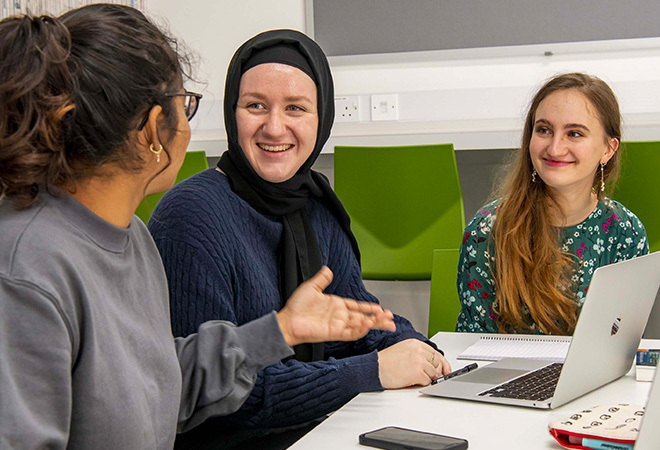 Three female students sat smiling and talking, the student in the foreground has her back to us and is talking to the other two students
