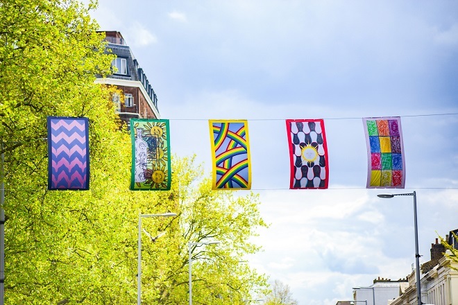 Photo of 5 different colourful flags with a tree to the left behind and a building