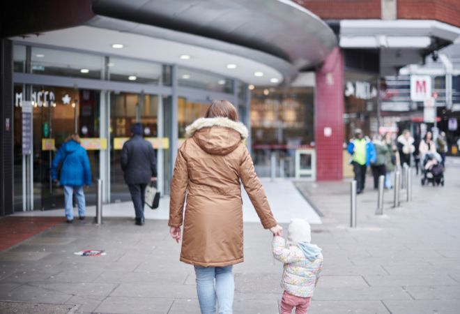 An adult, likely a parent, holding the hand of a small child as they walk towards the entrance of a building with a curved overhang and glass doors. The building has signage that reads “Morrisons.” There are several other individuals in the vicinity, walking or standing.