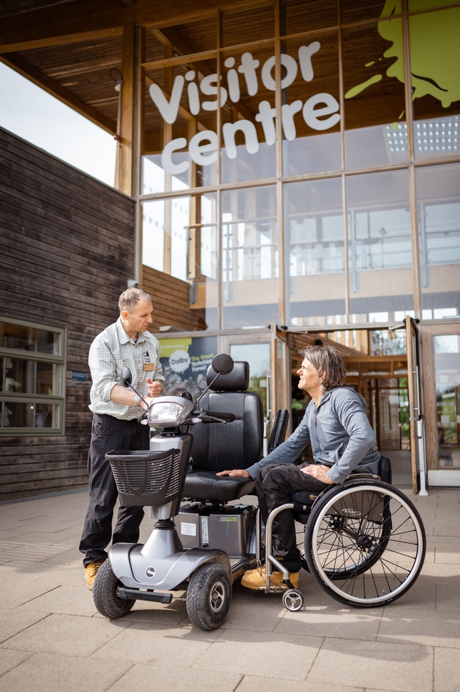 "Male wheelchair user talking to a male stood next to him both facing an electric powered wheelchair. Stood outside of a visitor centre in a forest"