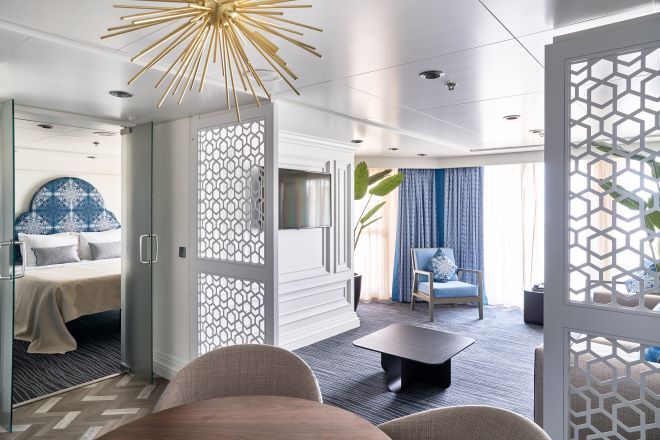 "A suite onboard Marella Voyager with a blue theme. There are double doors leading to the double bedroom, a dining table in the foreground and a living area behind sliding privacy screens. There is a TV, coffee table, blue chair and floor length curtains in the living area."