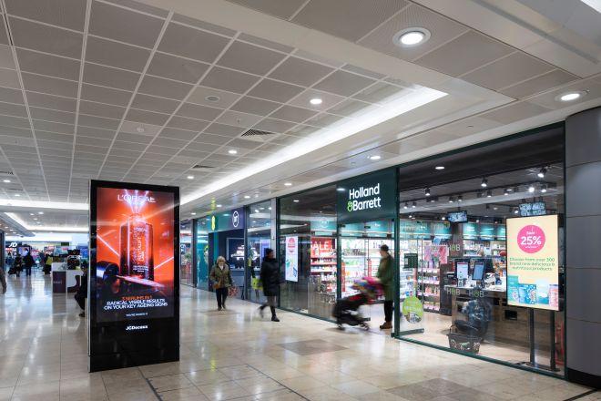 "Indoor shopping centre walkway with Holland & Barrett store and people walking past retail units."