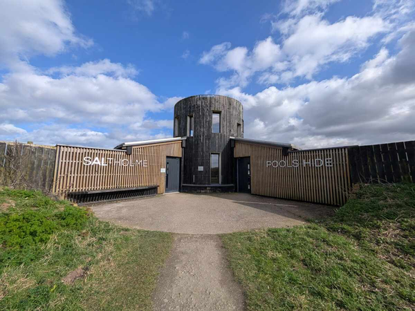 A circular wooden bird hide building with “Saltholme Pools Hide” signage stands at the end of a path under a partly cloudy sky