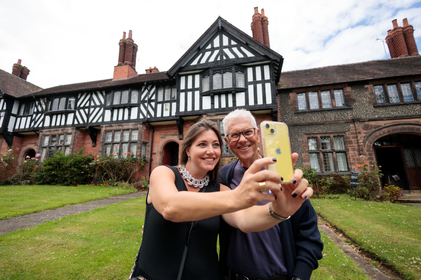 A woman wearing a black dress takes a selfie with a man in front of a large, picturesque, Tudor-style house and manicured lawn.