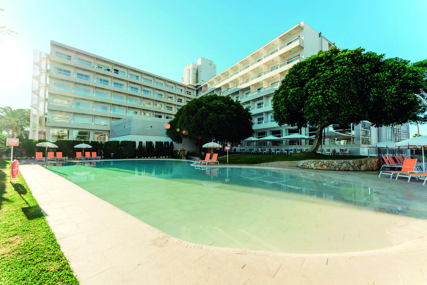 TUI Suneo Haiti hotel with some trees, orange sun loungers and umbrellas and the swimming pool in front of it.