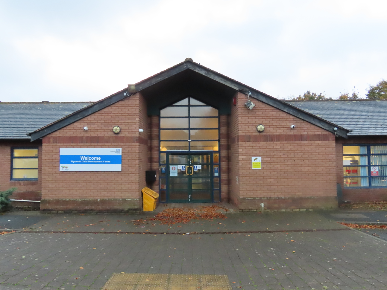 Exterior of Plymouth Child Development Centre, a single-story brick building with a pitched roof. The entrance features a large glass door and windows framed in dark trim. A sign reading 'Welcome Plymouth Child Development Centre' is displayed on the left side of the building. The surrounding pavement is lined with scattered autumn leaves.