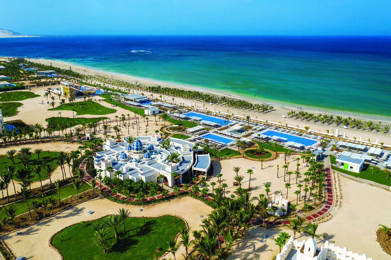 Aerial view of the large hotel complex with the hotel building in the middle surrounded by many palm trees, with pools right on the beach front. There are many, many sun loungers on the beach with the bluey green sea.