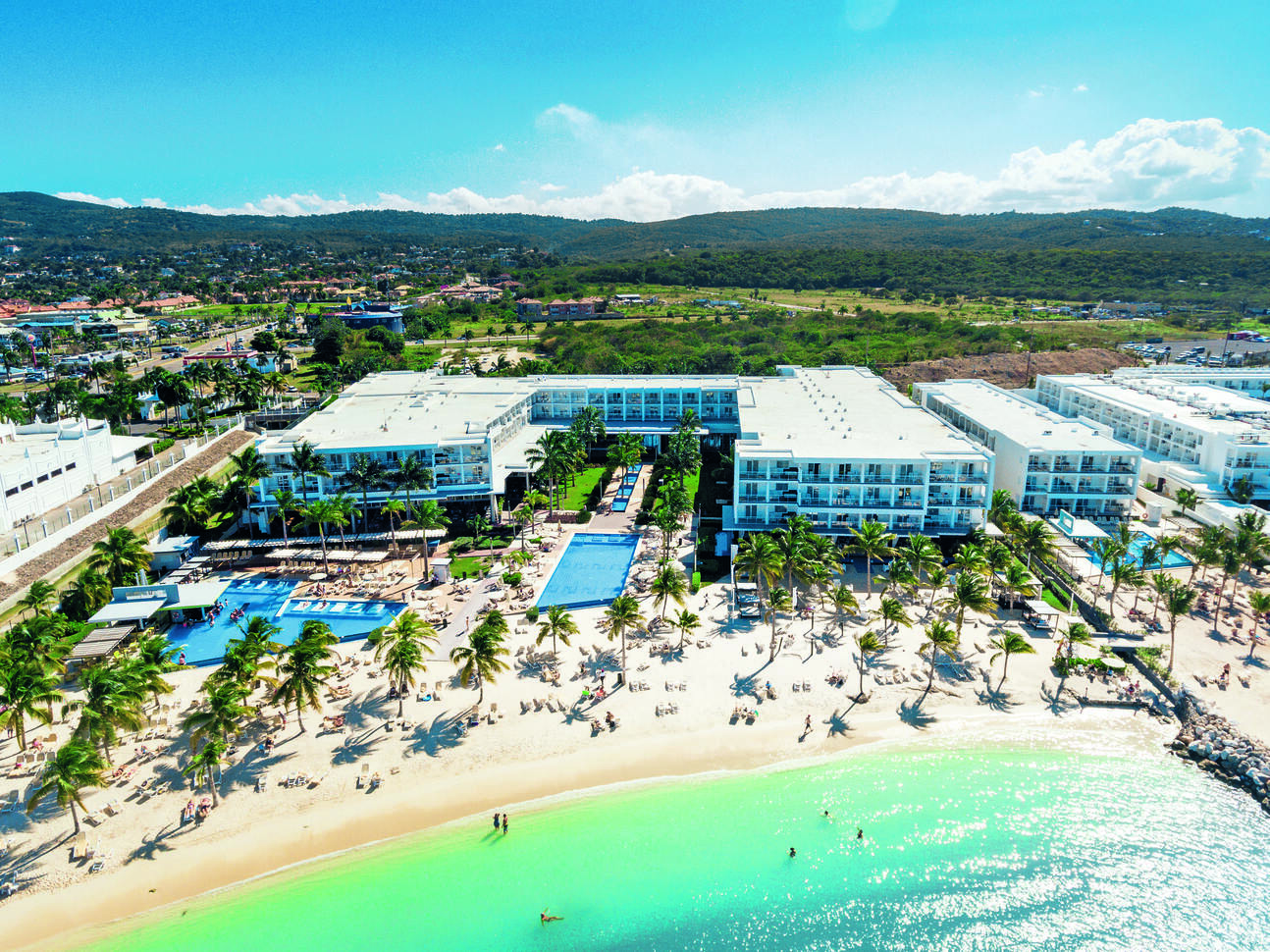 Aerial view above the green/blue sea and golden beach dotted in palm trees with the large, white hotel building behind with pools out the front.