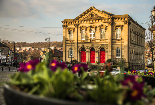 The front of Batley Methodist Church. A large stone building with 3 big red doors and many windows around the building. Close up flowers in the front of the image blurred out and the grey sky above.