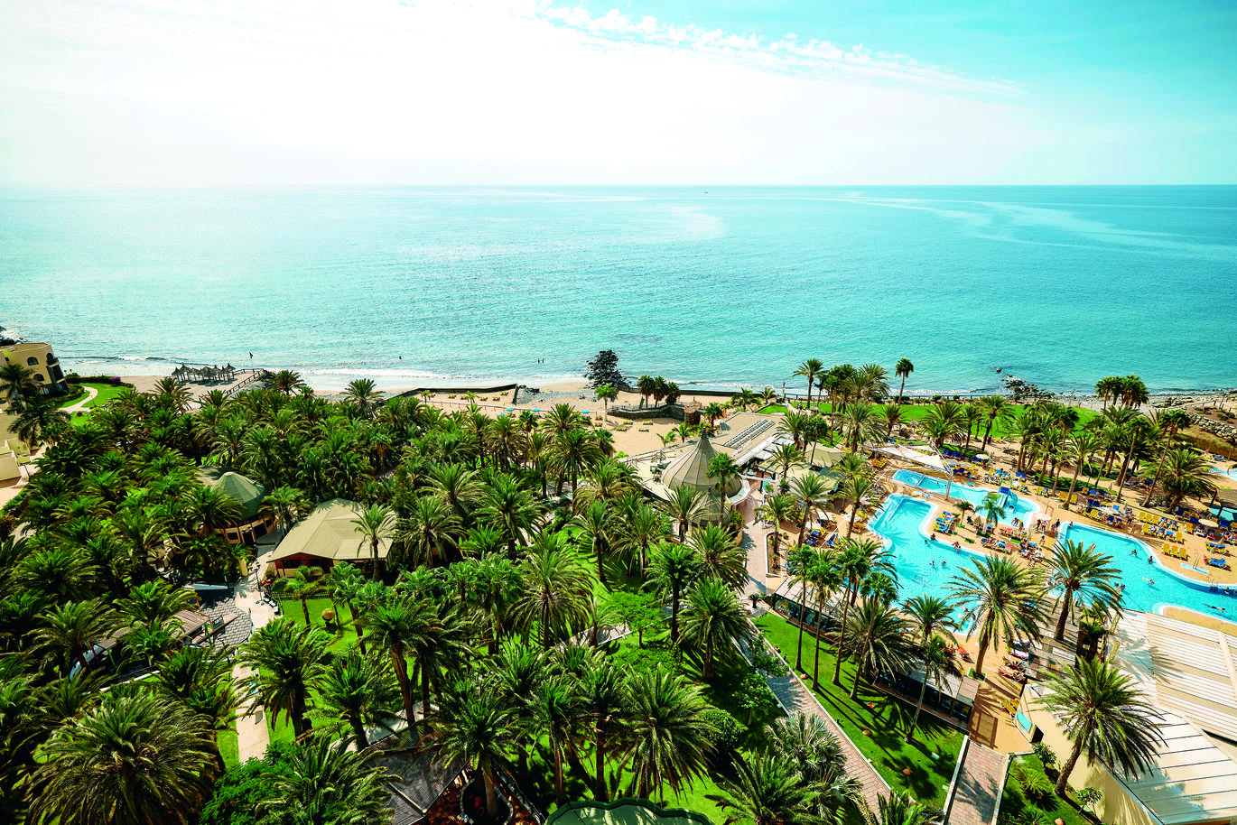 Aerial view of of the hotel pool surrounded by many many palm trees and the beach and lovely blue sea behind in the sun light.