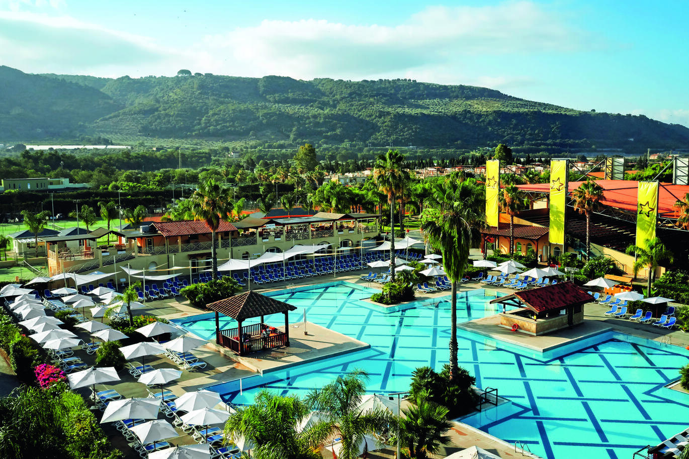Aerial view of the pool area surrounded with sun loungers and umbrellas with the resort surrounded by many trees and hills in the background.