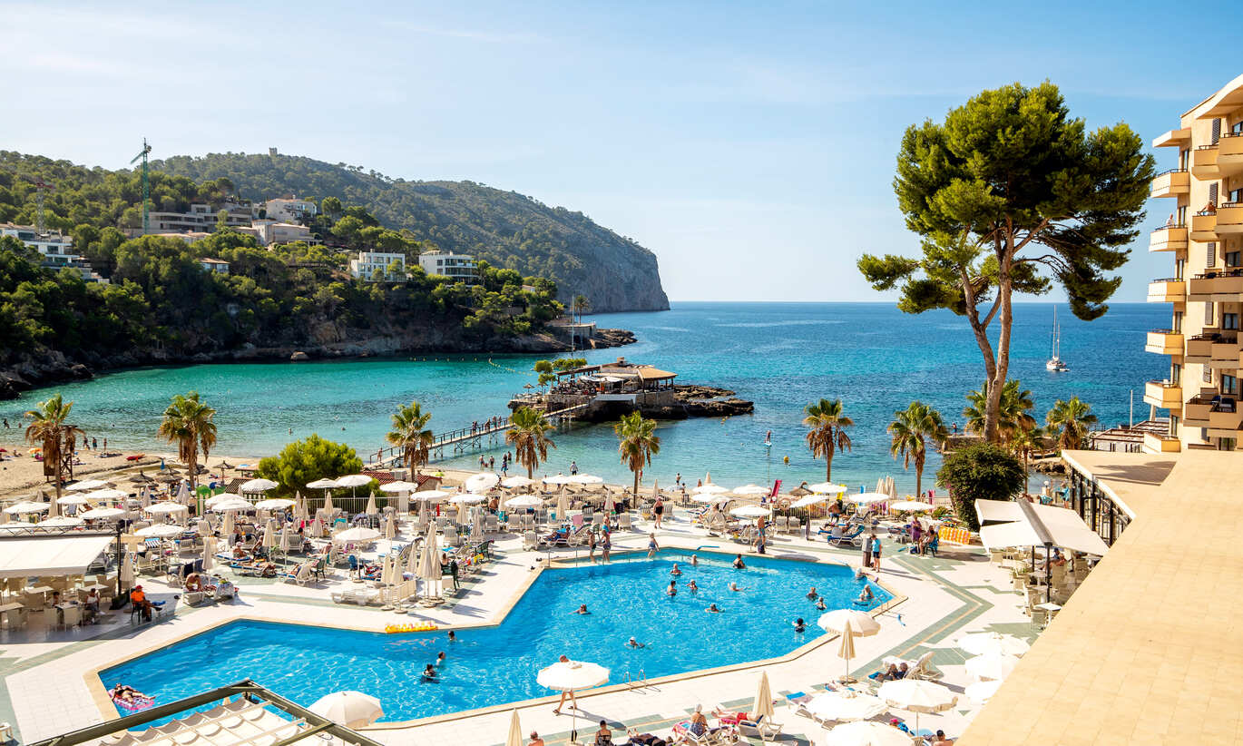 Picture taken from high up of the sea with hills behind with buildings and lots of greenery. There is a bridge from the beach leading out to a little hut on the sea. The pool is directly behind the beach with lots of sun loungers and umbrella's, lots of people around and in the pool. A few trees around the pool and a section of a cluster of rooms to the far right of picture. 