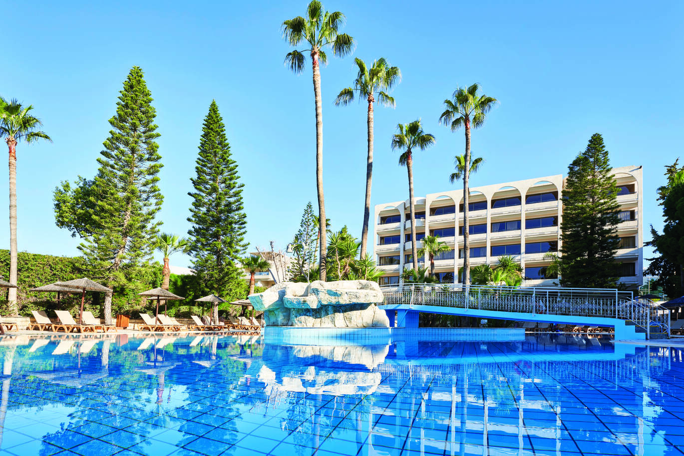 View from the side of the pool surrounded by sun loungers with straw umbrellas with many trees dotted around and a bridge over the pool. With a cluster of hotel rooms and balconies behind with the clear, blue sky above.