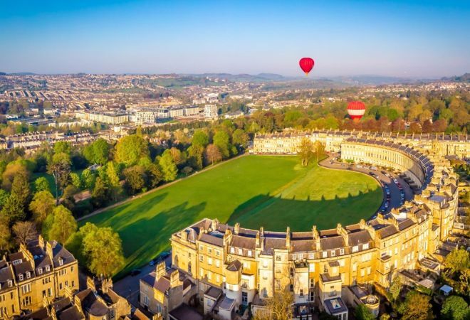 "Aerial image of Royal Crescent in Bath. A C shaped building with a large green on one side and hot air balloons in the background set against a blue sky and the backdrop of Bath"