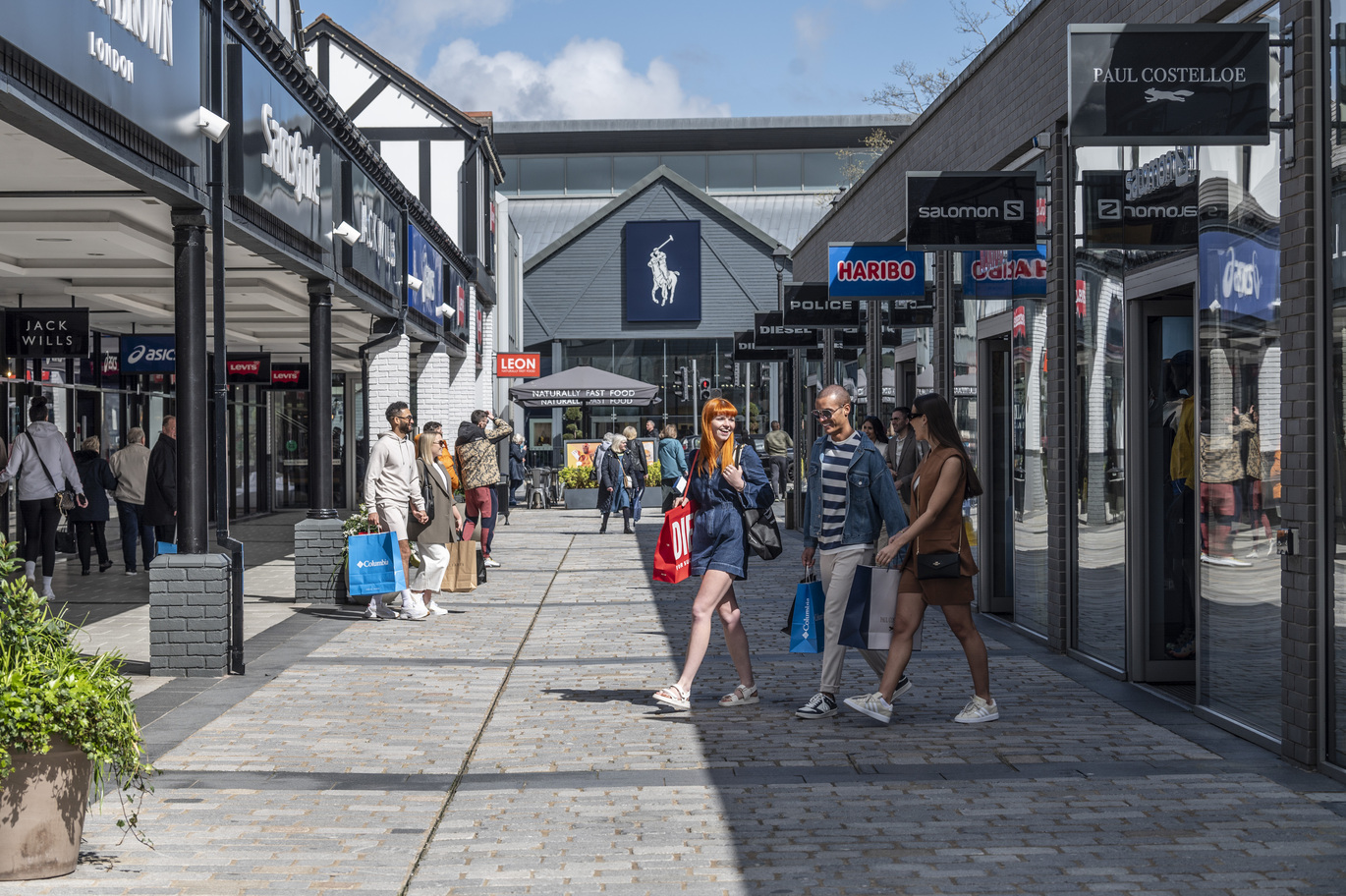 "People outside shops. Retail shops either side with a walkway down the middle"