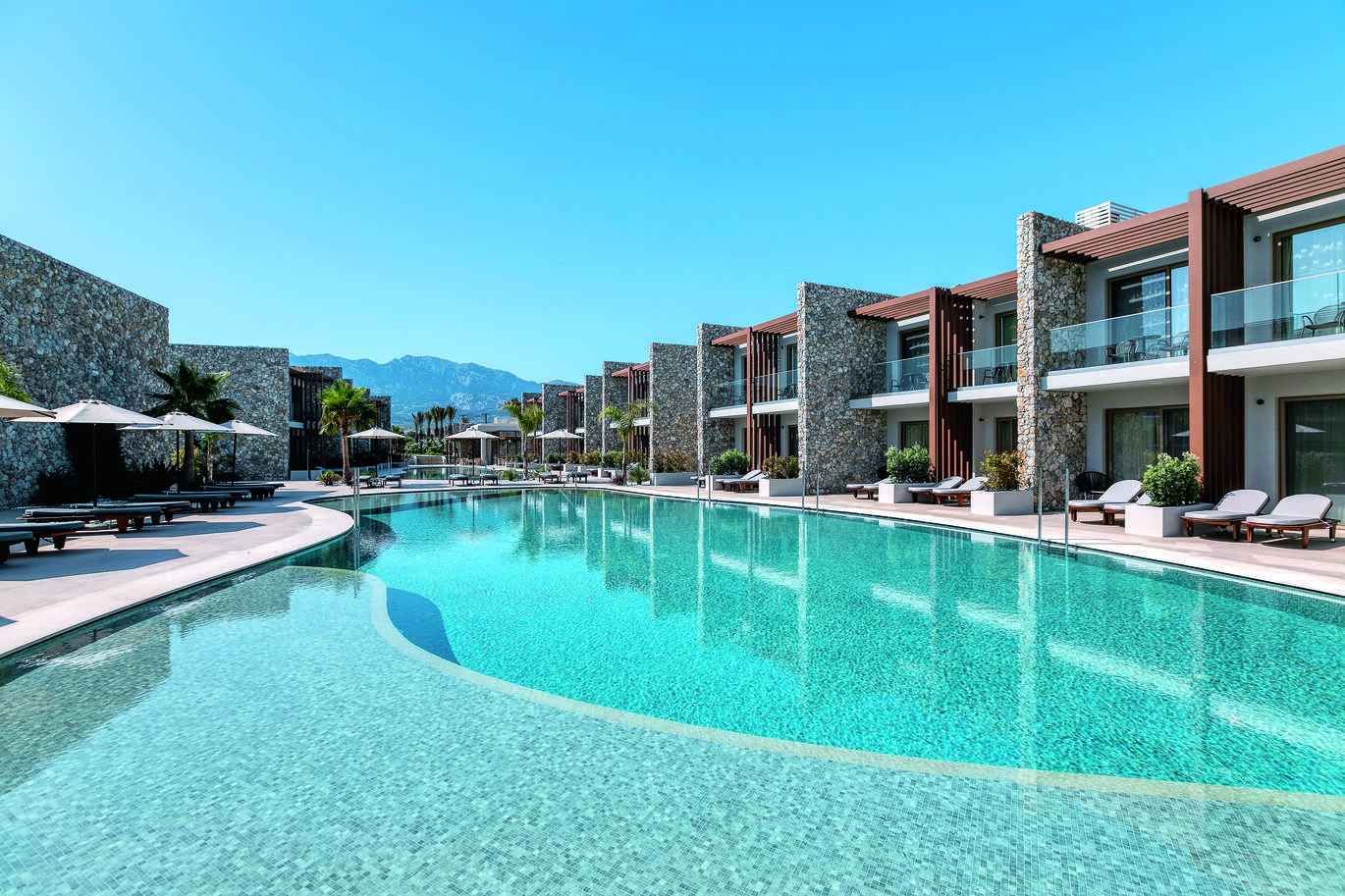 View of the pool area with sun loungers and umbrellas around the pool, clusters of hotel rooms to the right and mountains in the background on a clear, sunny day.