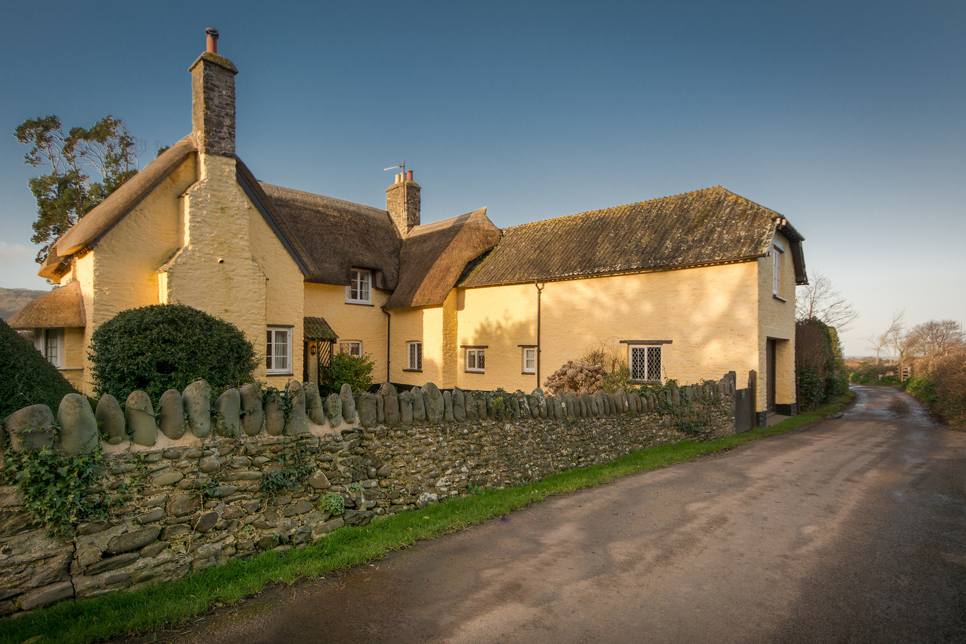 The exterior of the large yellow Holnicote Lower House with a stone wall out the front leading to the road.