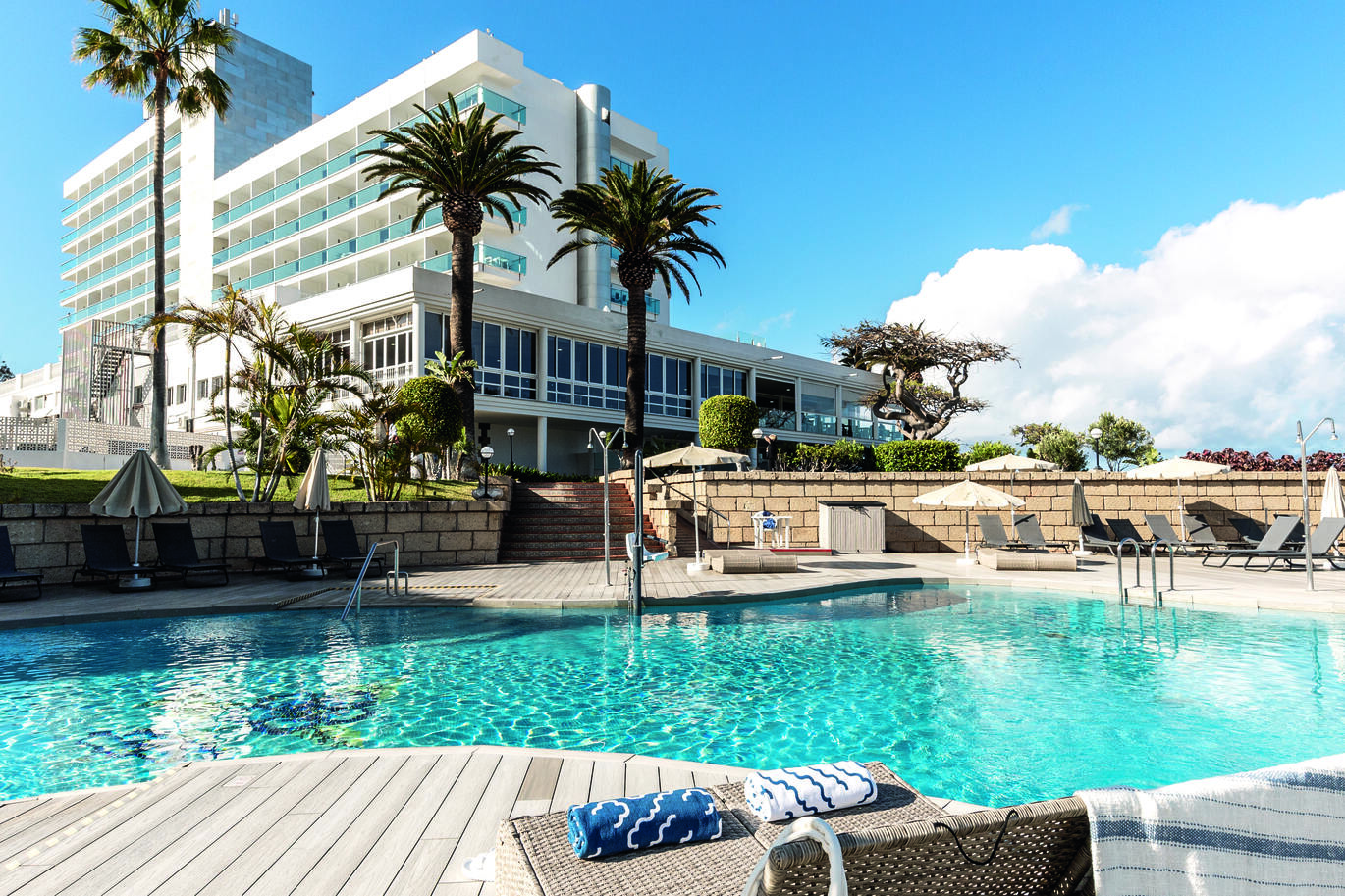 View from the pool area of the tall hotel building through some tall palm trees with the slightly cloudy but blue sky above.