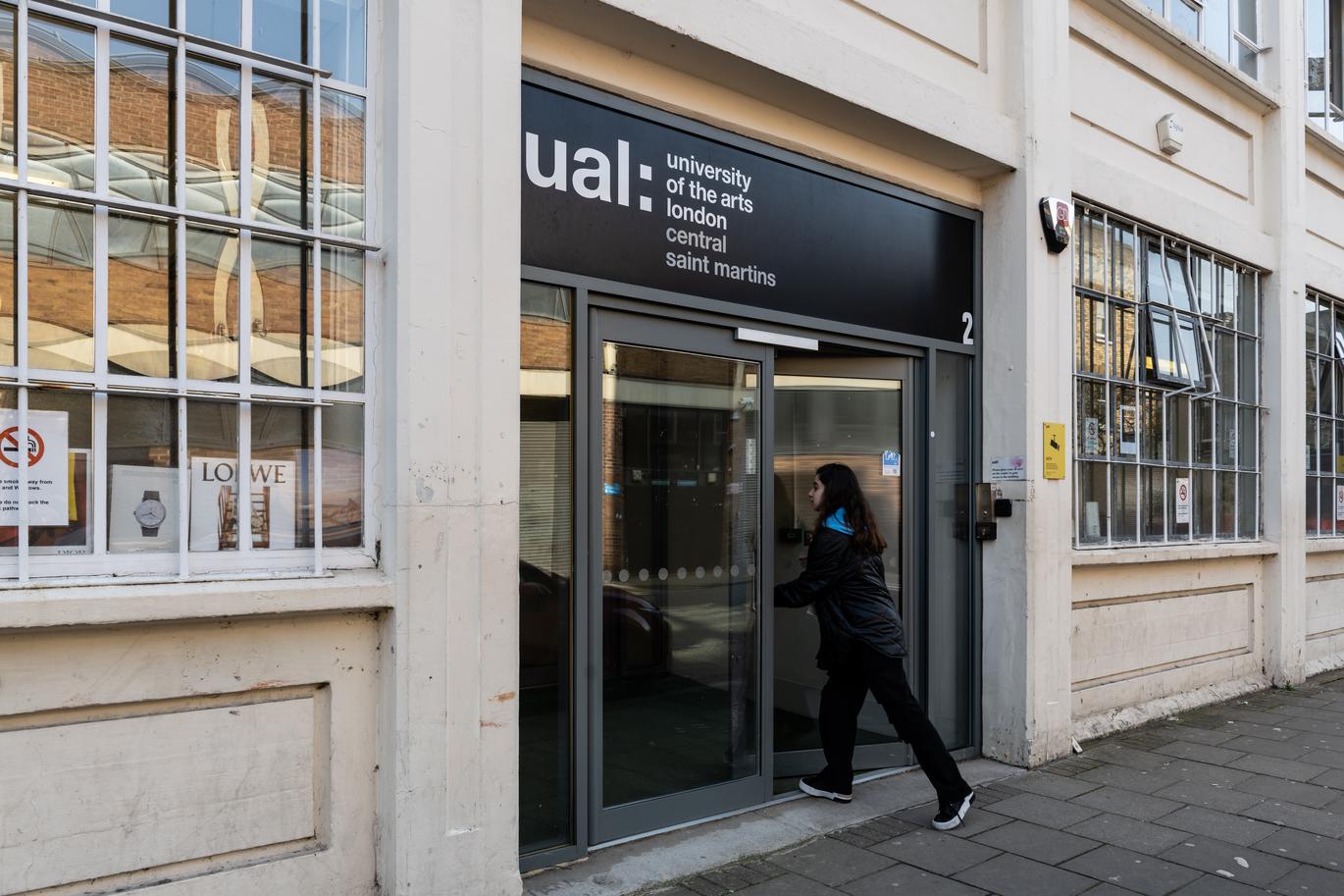 "Entrance to the UAL Central Saint Martins Archway building, showing a person pushing open a glass door beneath a black sign that reads “ual: university of the arts london central saint martins.” The exterior is cream-coloured with large paned windows and a paved walkway in front."