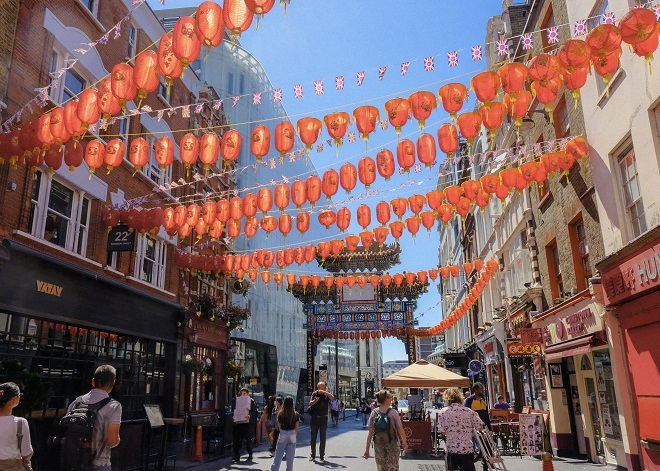 "People walking through Chinatown with shops and restaurants either side, large red lanterns streaming between the buildings above and a clear blue sunny sky."