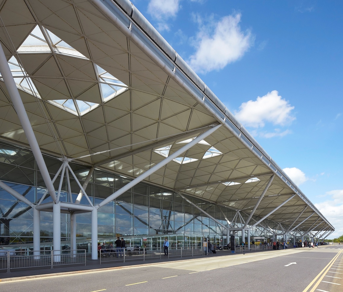 "External photo of Stansted Airport terminal building. This is the same as the hero image at the top of this page. There is parking to the right of the photo, a road in the middle and the building to the left. There is a large roof covering the path the full length of the building in the photo."