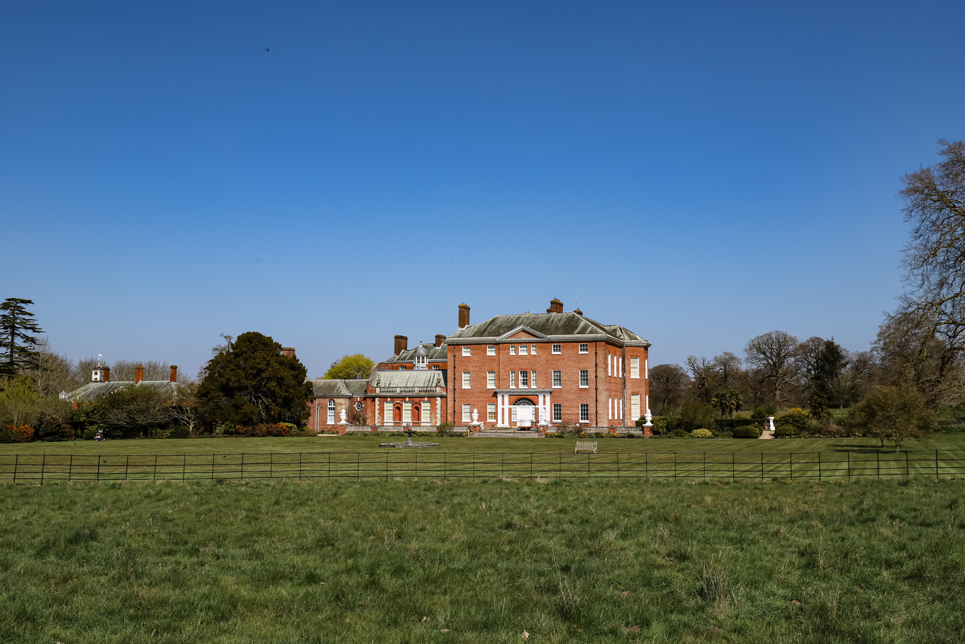 Exterior of the 18th century mansion with a large green lawn surrounded by small black fence, the mansion is surrounded by greenery and trees on a clear, bright day.