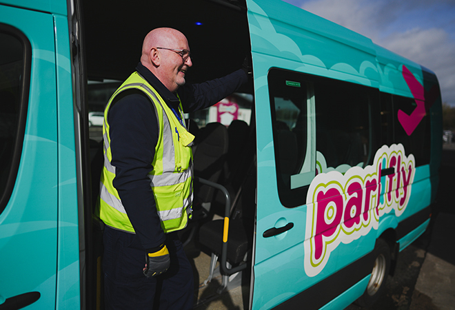 "Photograph of a turquoise park and fly bus with the driver, a white male wearing a fluorescent waistcoat stood in the doorway"