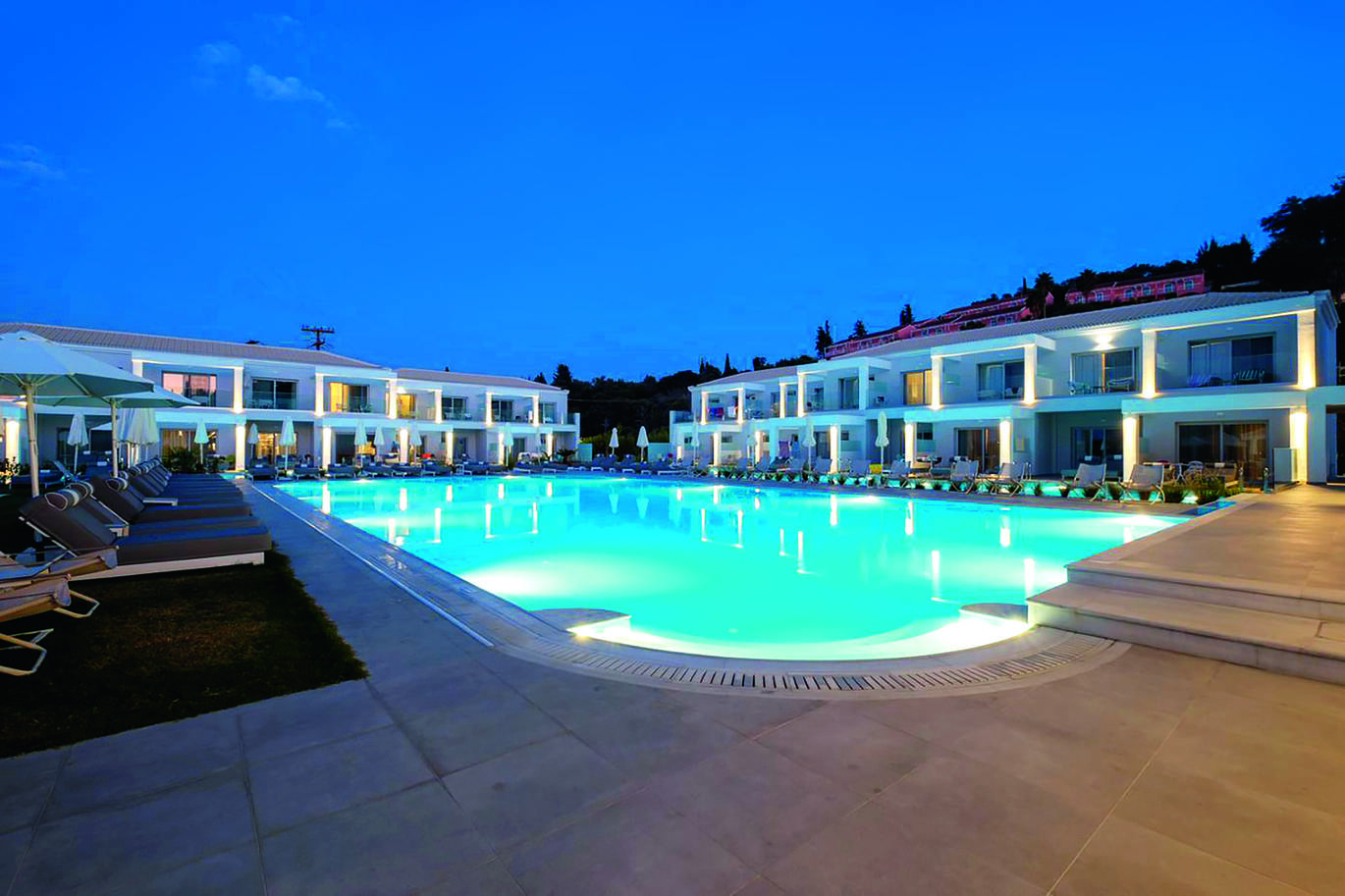 Dusky view of the pool area with sun loungers and umbrellas surrounding and the hotel buildings behind.
