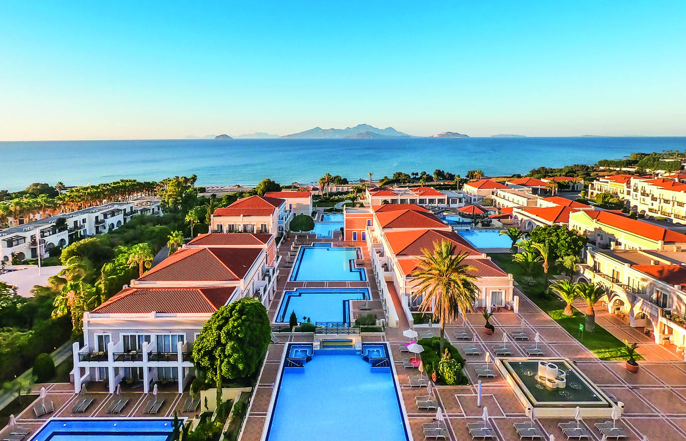 Aerial view of the hotel complex with the pools in the middle of the buildings and the sea behind with mountains in the distance.