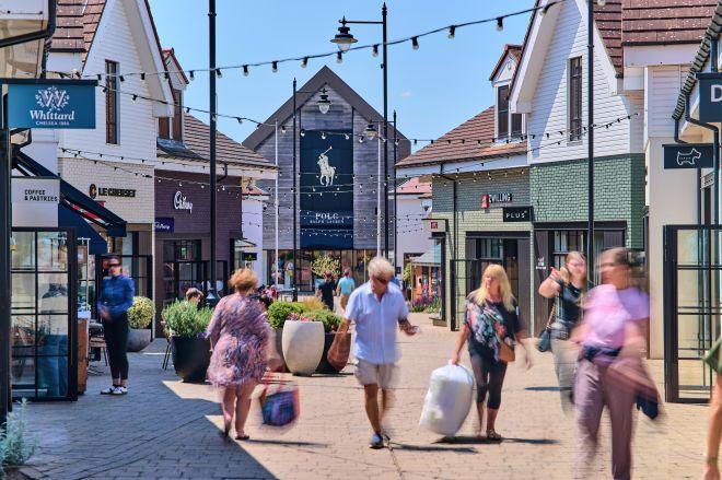 "Shoppers walk along a pedestrian street at Braintree Village, lined with outlet stores and string lights overhead."