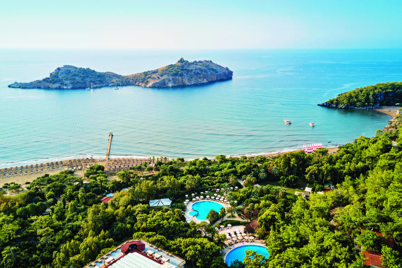 Aerial view of the pool and beach area surrounded by a forest of trees with 2 pools visible in the middle with the beach and sea behind and a small island not far from the beach. A few small boats are also seen in the sea.