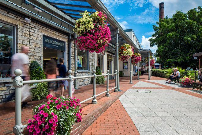"Clarks Village walkway with hanging flower baskets, stone-fronted shops and visitors walking along the path."