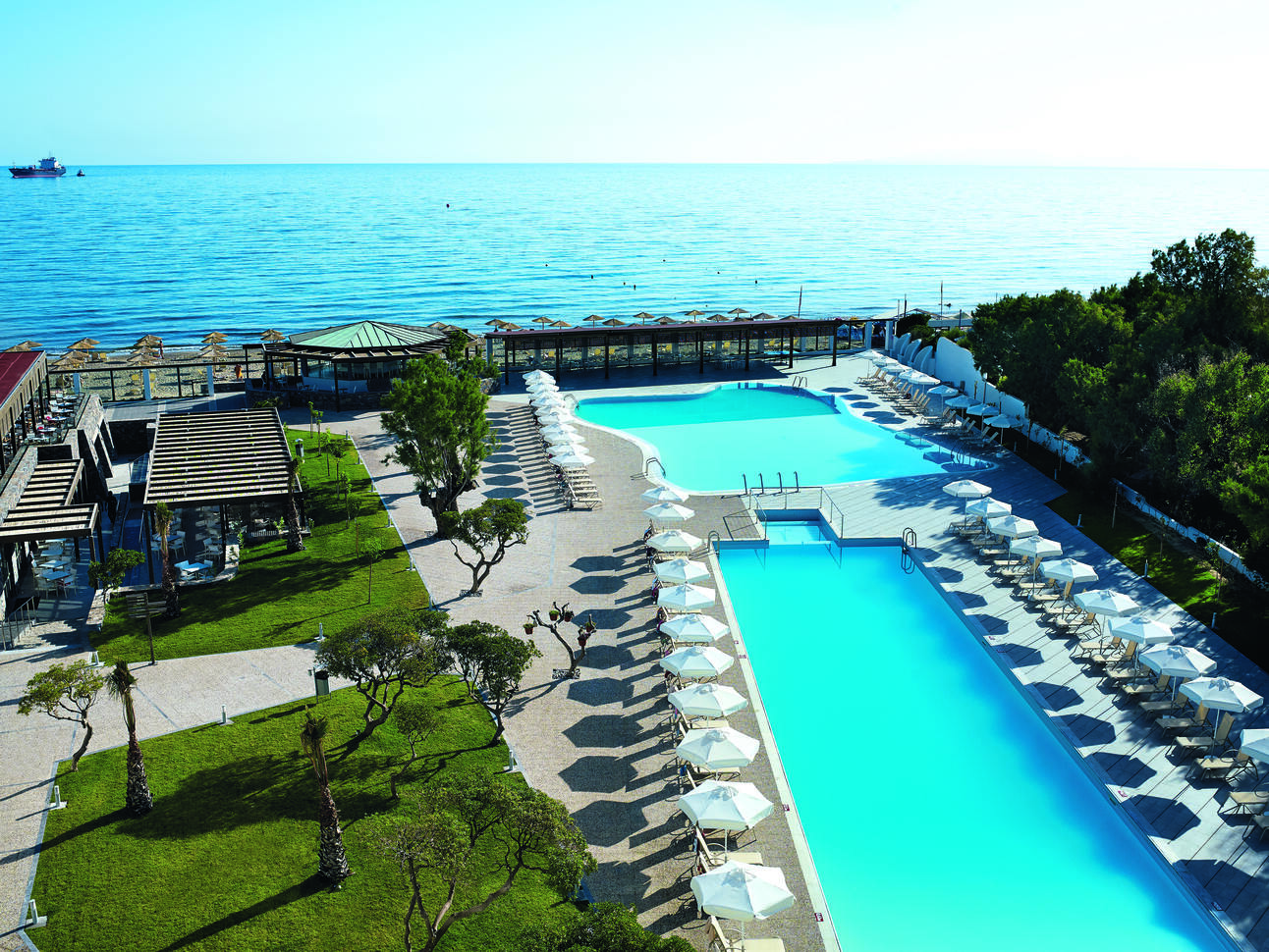 A long clear blue pool and a smaller blue pool surrounded by white sun loungers and umbrellas with a lawn, trees and a couple of shelters/huts around the pool area. The beach is directly behind with more sun loungers and umbrellas and the lovely blue sea with a large boat in the back ground. The sky above is clear and blue.