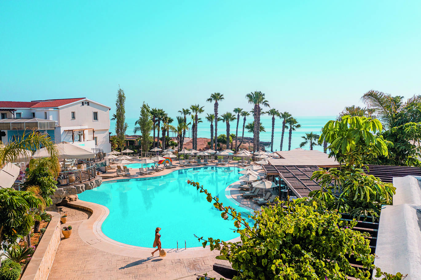 Aerial view of the pool area with the sea behind through the palm trees, with the hotel buildings surrounding the pool and surrounded by deck chairs and umbrellas on a clear, sunny day.
