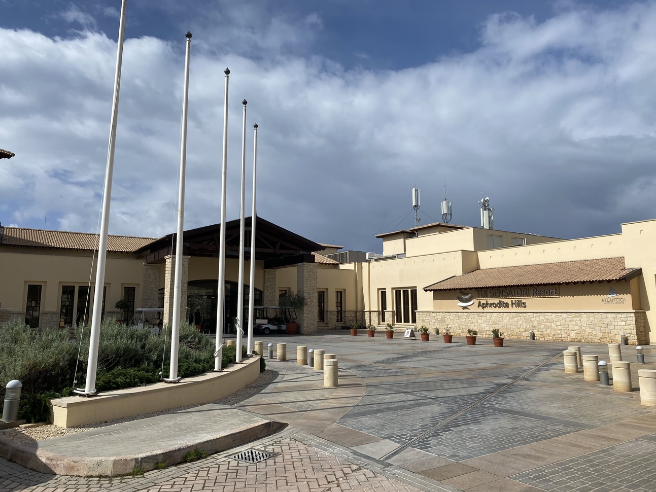 TUI BLUE Sensatori Atlantica Aphrodite Hills front of building, sand colour with brick flooring, flower pots leading up to the entrance and some large white poles to the left.
