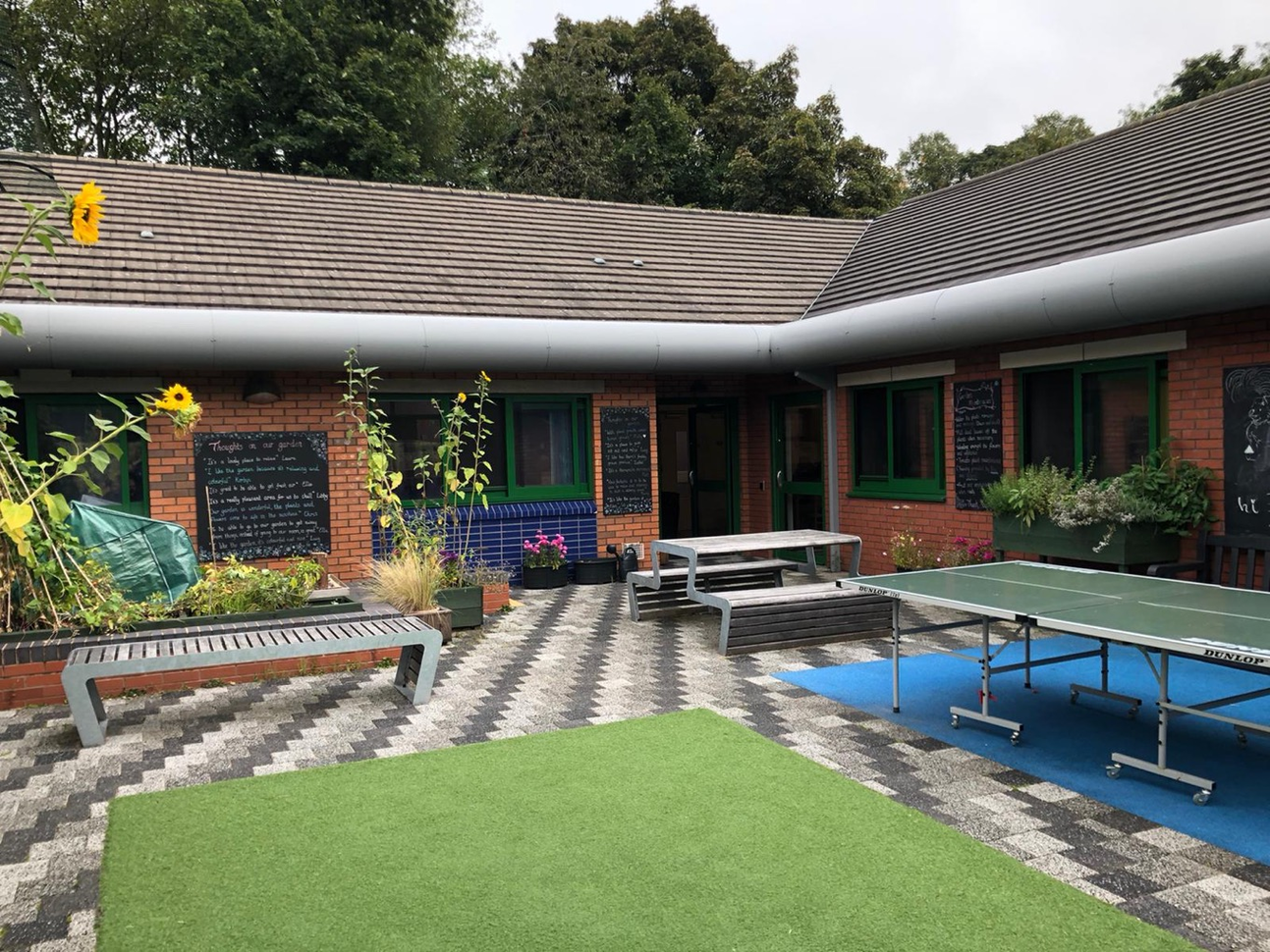 Courtyard with a ping pong table. Some black boards on the wall with "thoughts on our garden" written in chalk. Court has lots of plants, seating and a table.