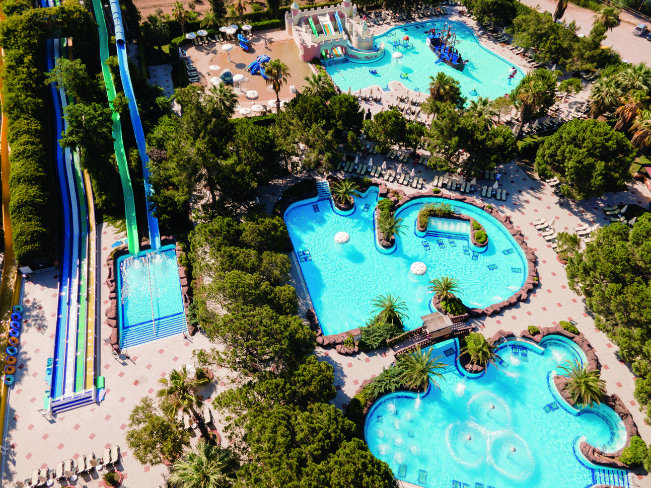Birdseye view of the pool area with 3 pools surrounded by sunbeds and trees, one pool with a pirate ship, slides and play equipment in and large water slides to the left of the pools.